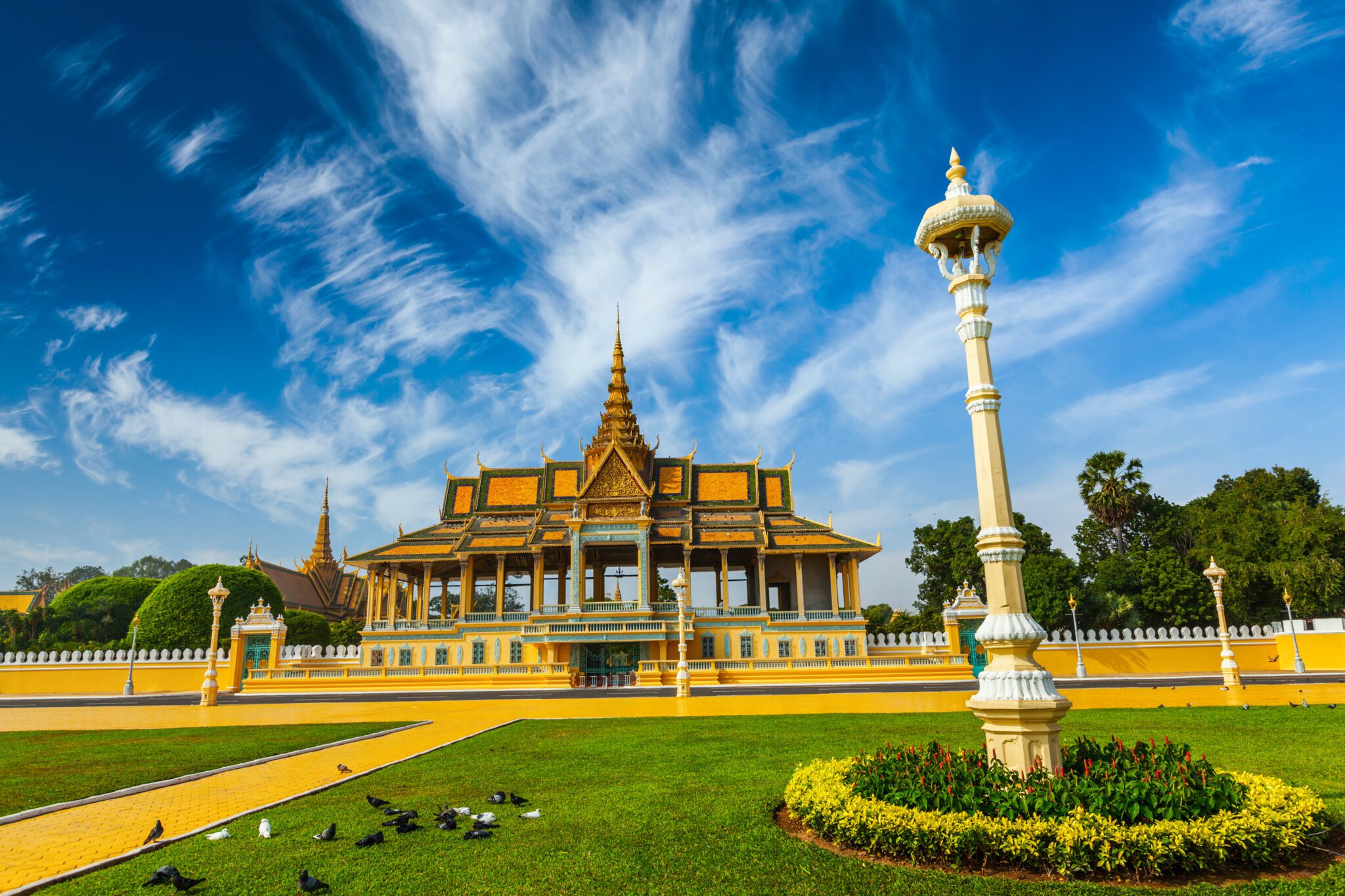 The Throne Hall and central structures of the Royal Palace in Phnom Penh, Cambodia, featuring traditional golden Khmer architecture, set against a bright blue sky and green lawn.