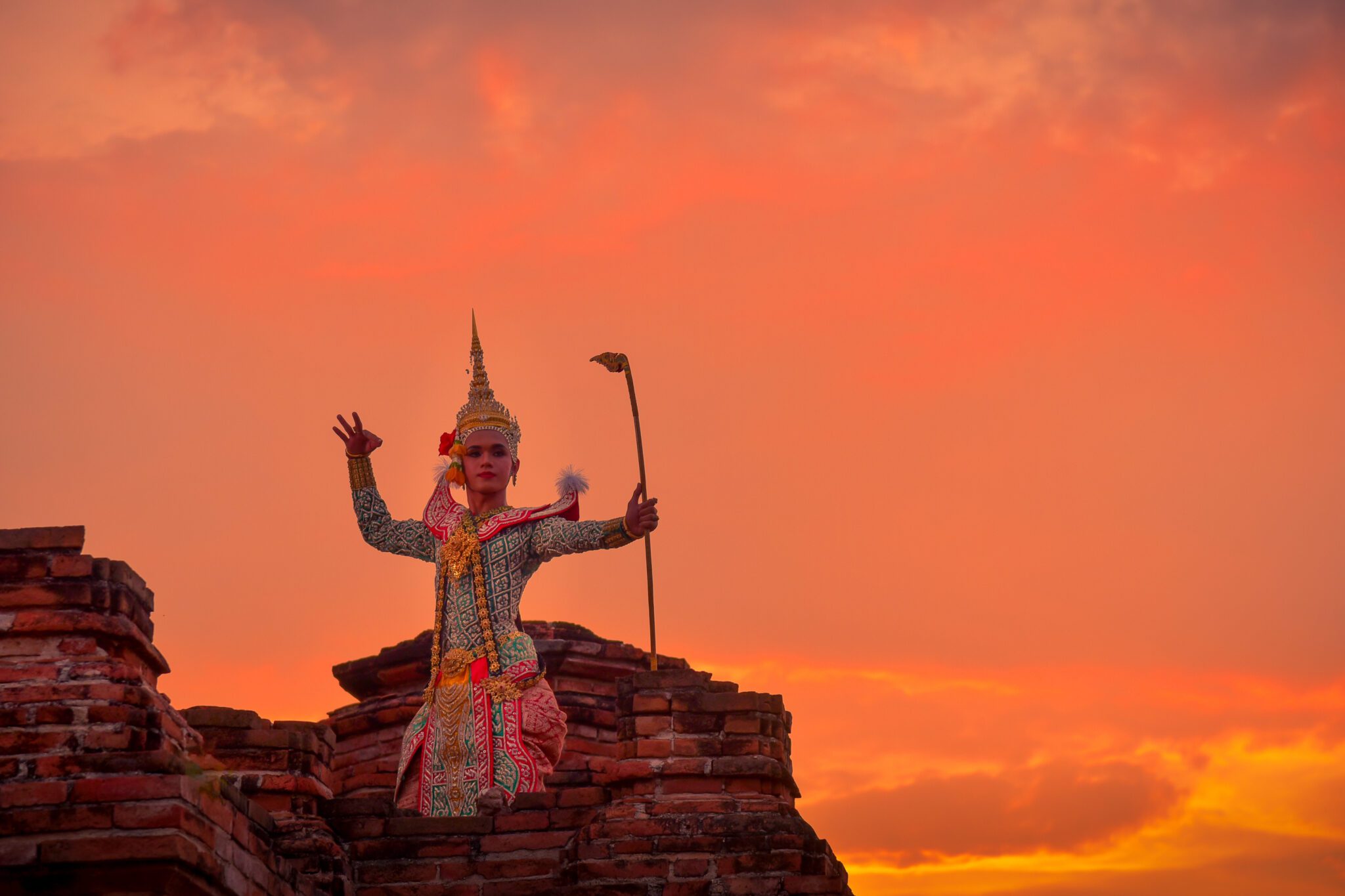 Traditional Khmer Apsara or classical dancer in elaborate gold costume and towering headpiece, posing on ancient red brick ruins against a dramatic sunset backdrop.