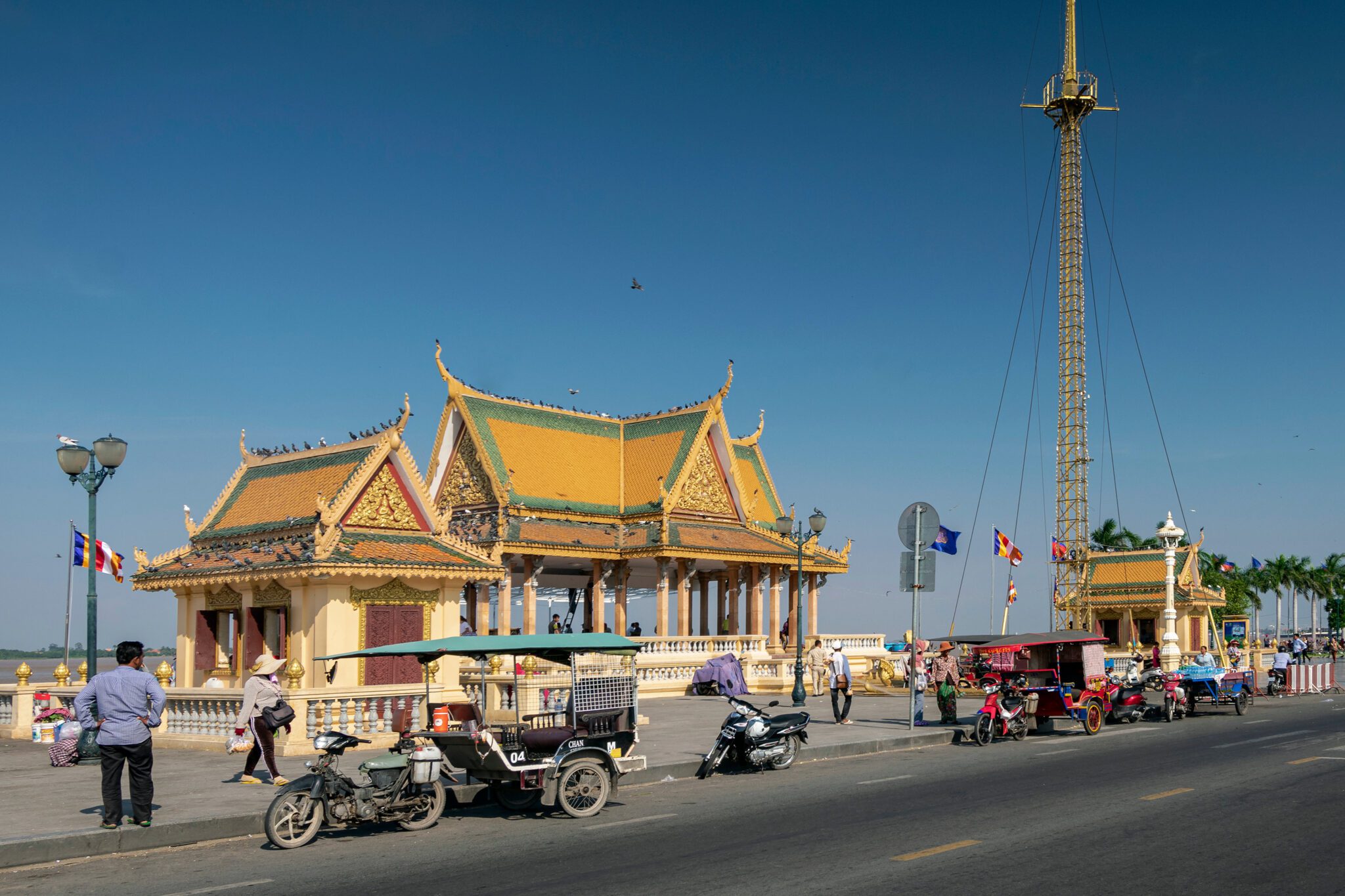 Traditional Khmer pavilion with a golden-yellow roof along a riverside promenade in Cambodia, with tuk-tuks and motorbikes parked nearby under a clear blue sky.