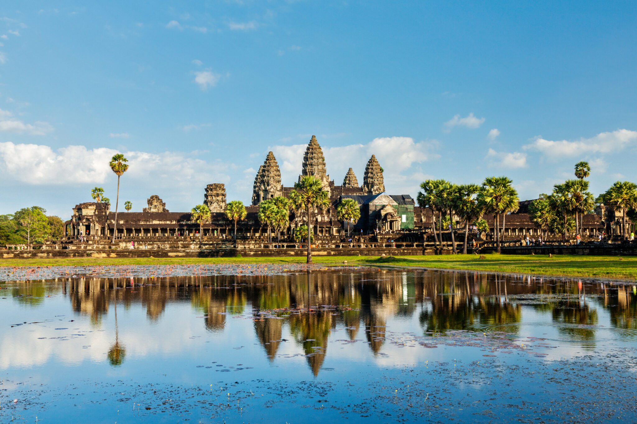 Angkor Wat temple complex in Siem Reap, Cambodia, reflected in a large pool of water in the foreground, with palm trees and a blue sky overhead.
