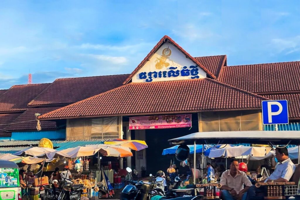 Busy local market with a large tiled roof, featuring a Khmer sign and stalls selling goods outside, with tuk-tuks and motorbikes parked on the street.