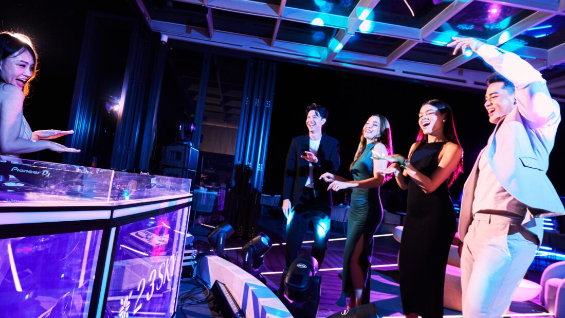 A female DJ mixing music on Pioneer decks with purple lights, while a group of happy, well-dressed people dance and celebrate on the 23 SKY rooftop bar.