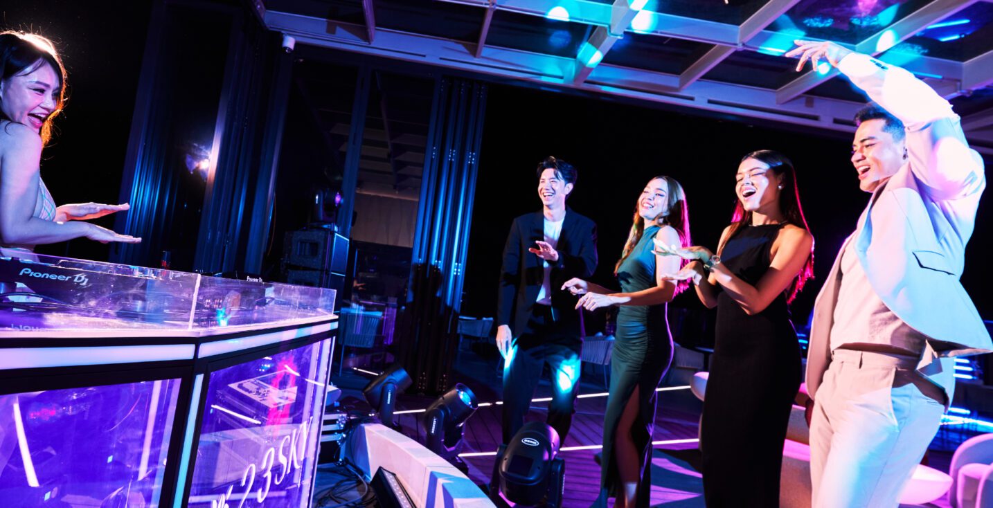 A female DJ mixing music on Pioneer decks with purple lights, while a group of happy, well-dressed people dance and celebrate on the 23 SKY rooftop bar.