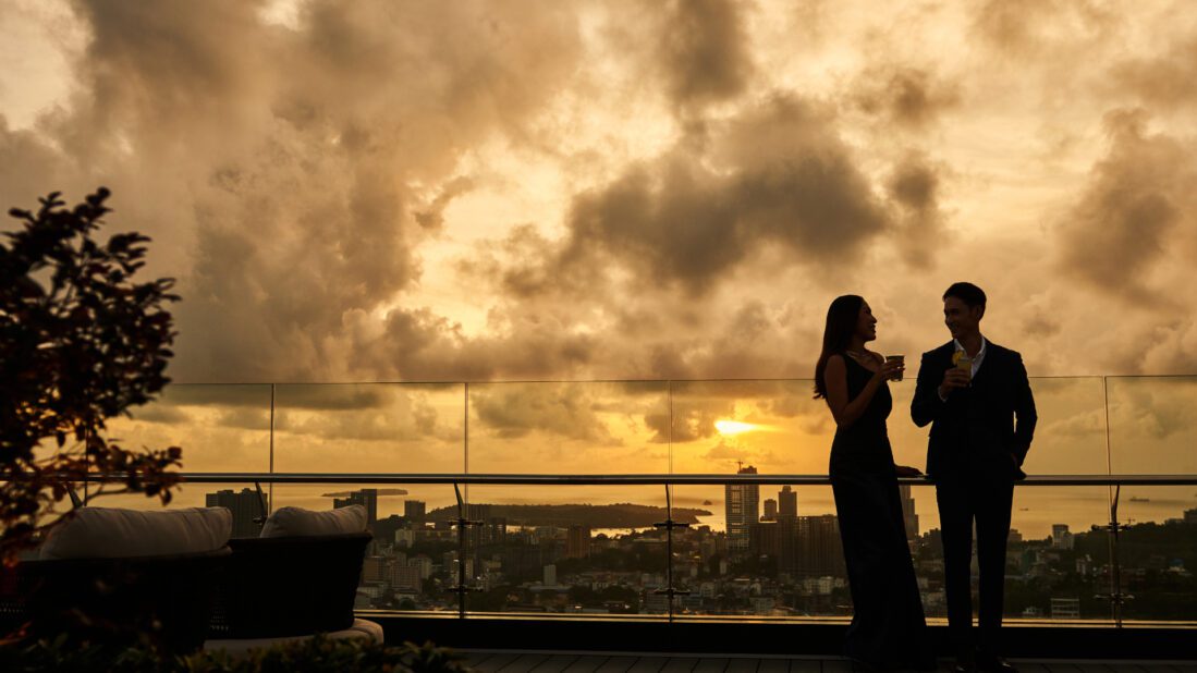Silhouette of a sophisticated couple enjoying cocktails on the 23 SKY rooftop terrace at Trimulia Hotel, overlooking a cityscape and ocean view beneath a dramatic sunset sky.