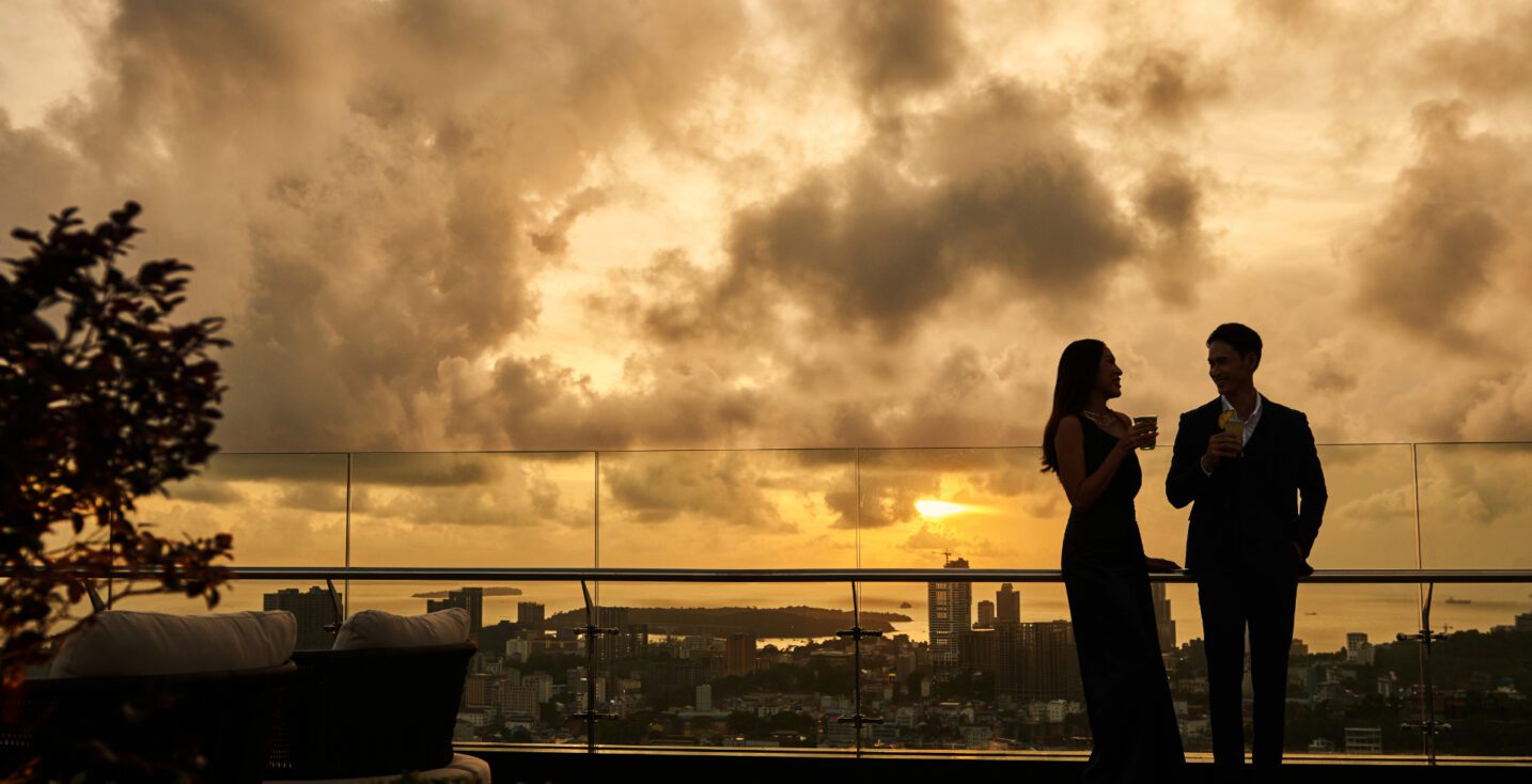 Silhouette of a sophisticated couple enjoying cocktails on the 23 SKY rooftop terrace at Trimulia Hotel, overlooking a cityscape and ocean view beneath a dramatic sunset sky.