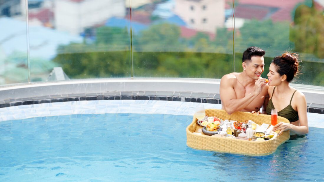 Close-up of a happy couple enjoying a breakfast served on a large floating tray in the Trimulia Hotel's rooftop infinity pool, promoting the luxurious 'Sweet and Sweat' experience.