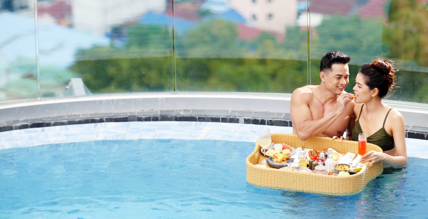Close-up of a happy couple enjoying a breakfast served on a large floating tray in the Trimulia Hotel's rooftop infinity pool, promoting the luxurious 'Sweet and Sweat' experience.